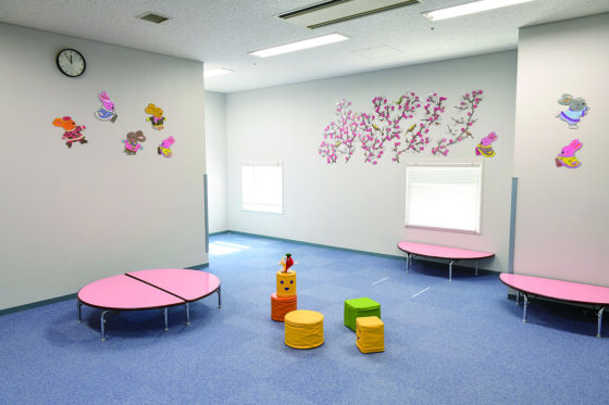 託児室の様子。青いカーペットの床にはピンクの円テーブルや丸や四角のオモチャが置いてある。オモチャの上にはミュートン。白い壁には8匹のウサギ、満開の紅白の梅の木が貼られている。