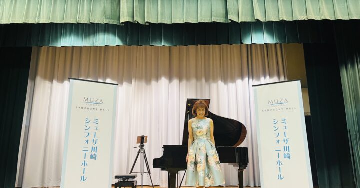小川典子さんがステージ上のグランドピアノの前でミューザのバナーに挟まれ、笑顔で立っている写真。衣装は鮮やかな水色に大きな花柄。