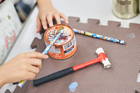 マットの上に置かれたさばの味噌煮缶を鉛筆で叩く子どもの手。マットには小さなハンマーも置かれている。